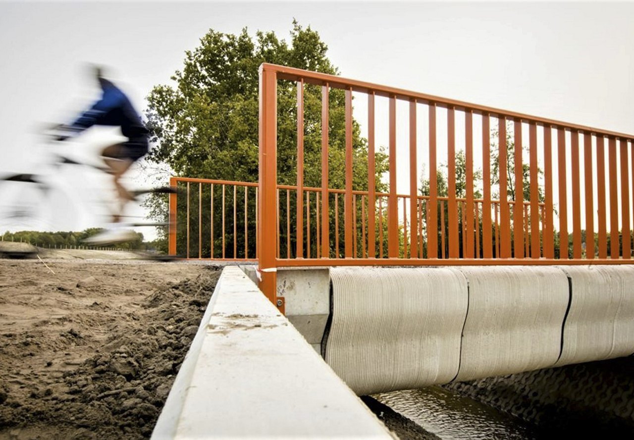 A cyclist in motion blur crossing a newly constructed 3D-printed concrete bridge with orange railings.