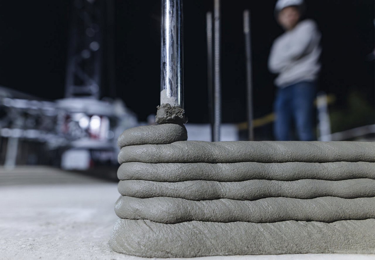 A building is being constructed using 3D printing technology, as cement is layered to form the base of the structure, with a worker standing behind it, monitoring the progress.