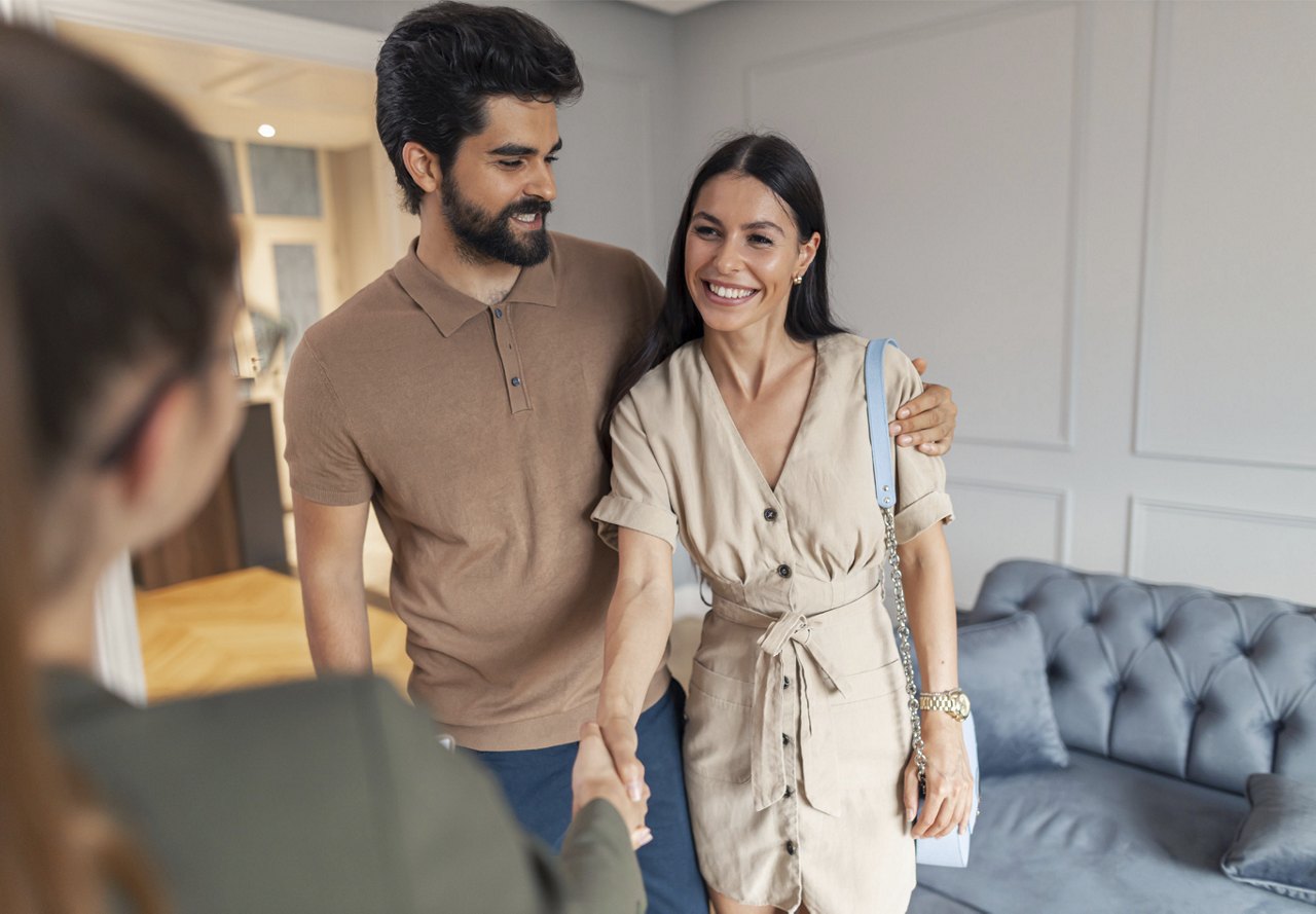 A smiling couple being greeted by someone in a modern living room, suggesting a friendly meeting or housewarming.
