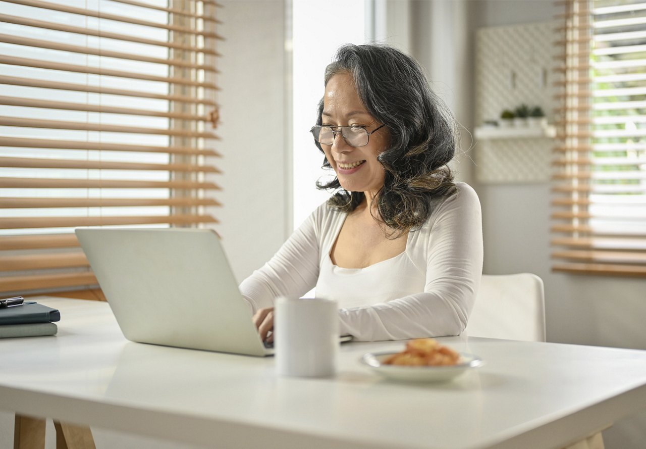 A cheerful mature woman working on a laptop in a bright, homey kitchen, with a cup of coffee and a plate of cookies nearby.