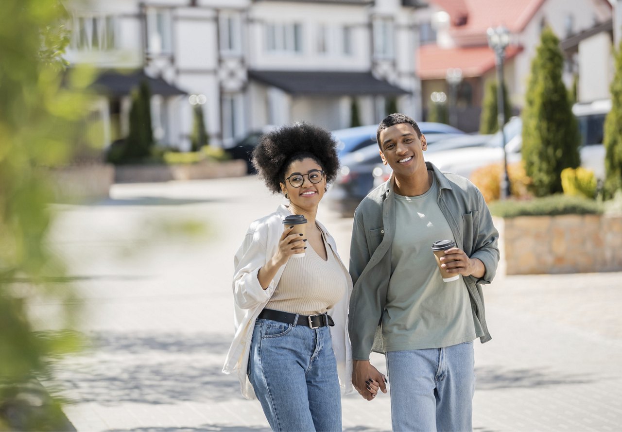 A happy young couple enjoying a walk in a residential neighborhood with coffee cups in hand.