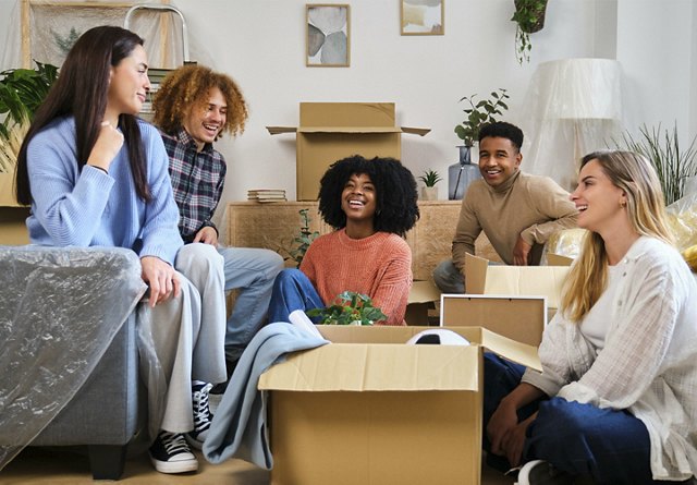 Four friends laughing together in a living room with moving boxes, indicating a moving day or new accommodation.