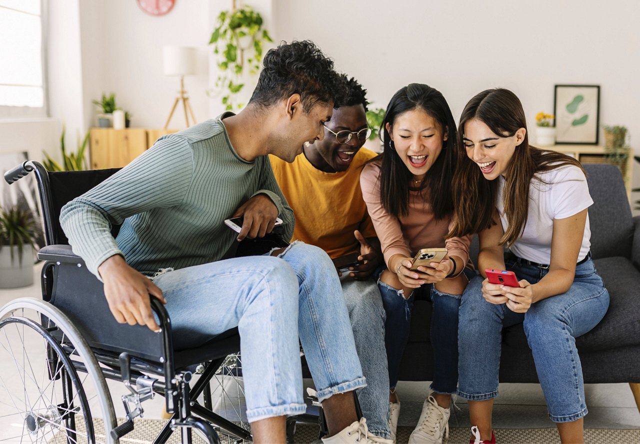 Group of four diverse friends, one in a wheelchair, laughing and sharing content on their smartphones.