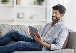Man sitting on couch in apartment looking at tablet with bookcase in the background