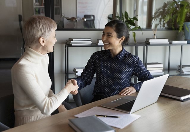 Two women shaking hands while sitting at table in front of computer