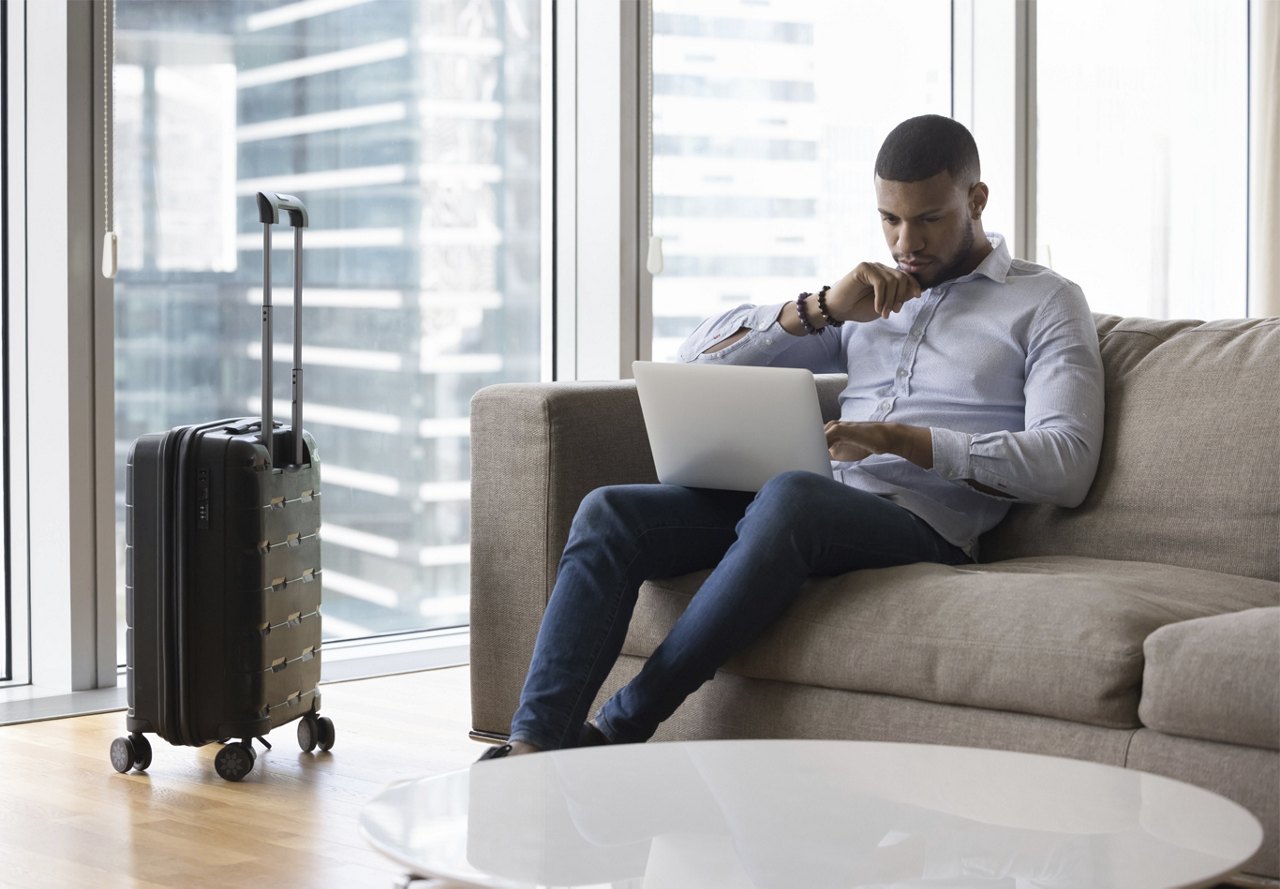 Man sitting on couch in apartment and working on computer with suitcase nearby