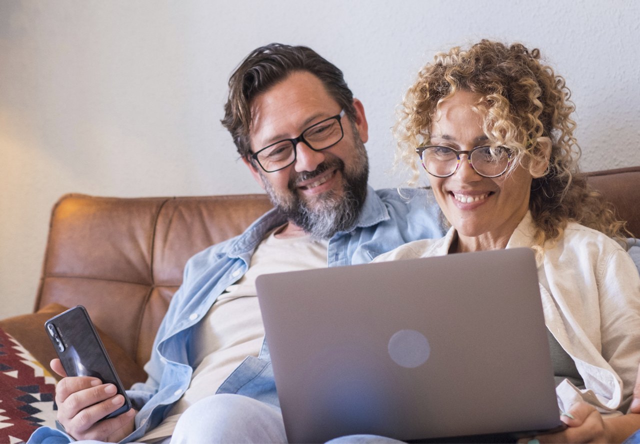 Older couple looking at computer screen smiling while holding cell phone