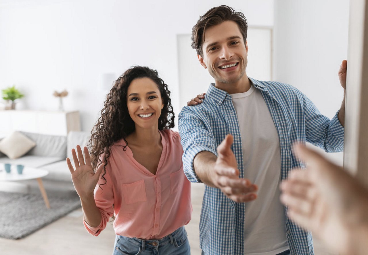 A young woman and man standing at their apartment door, warmly greeting a visitor with a wave and an extended hand for a handshake, ready to welcome them inside.