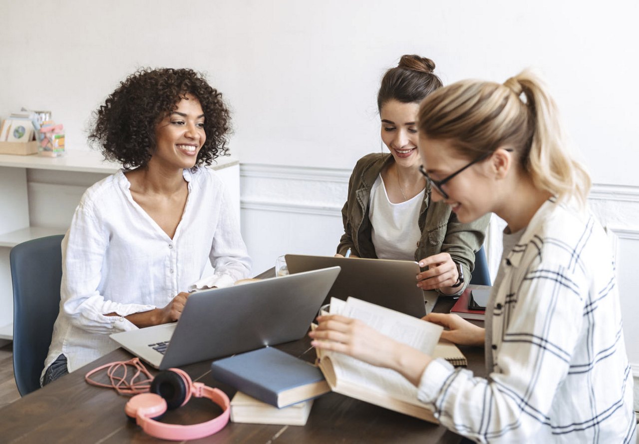 Three young women sitting at a table in a student housing common area, surrounded by laptops and books, focused on studying together in a collaborative and cozy environment.