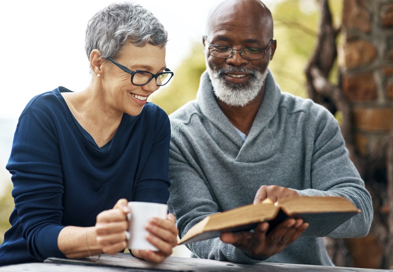 Smiling mature couple enjoying a book together outdoors, with the woman holding a mug.