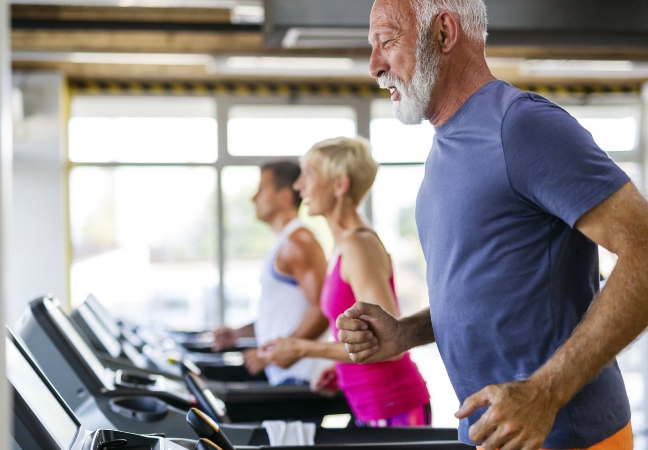Elderly man with a white beard running on a treadmill in the foreground, with more people exercising in the background.