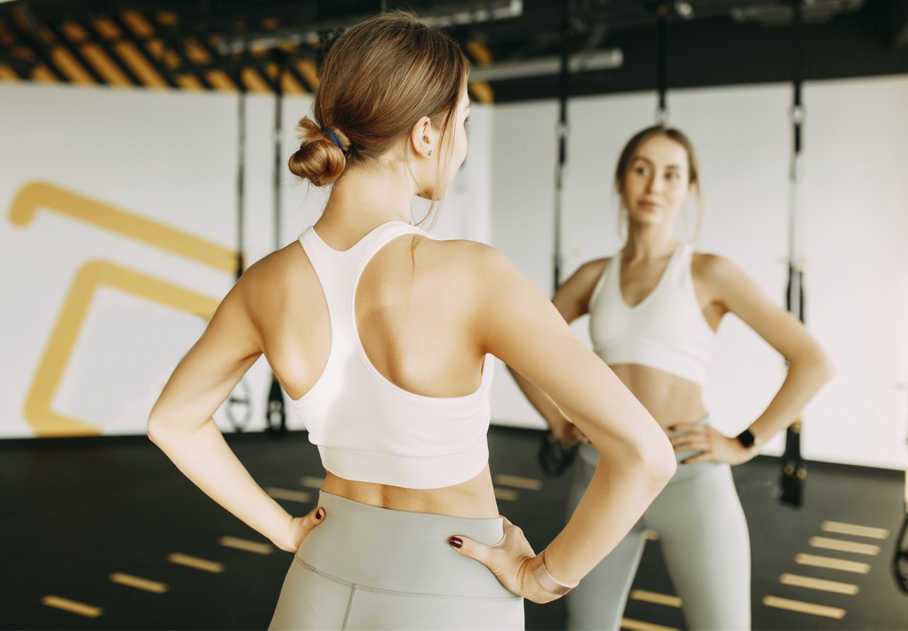 A woman in a white workout top and grey leggings stands facing a mirror, hands on hips, in a brightly lit gym.