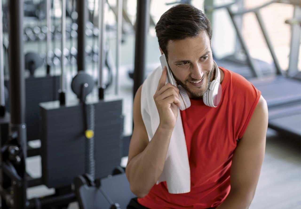 "Smiling man in a red tank top with headphones around his neck, taking a phone call in the gym.