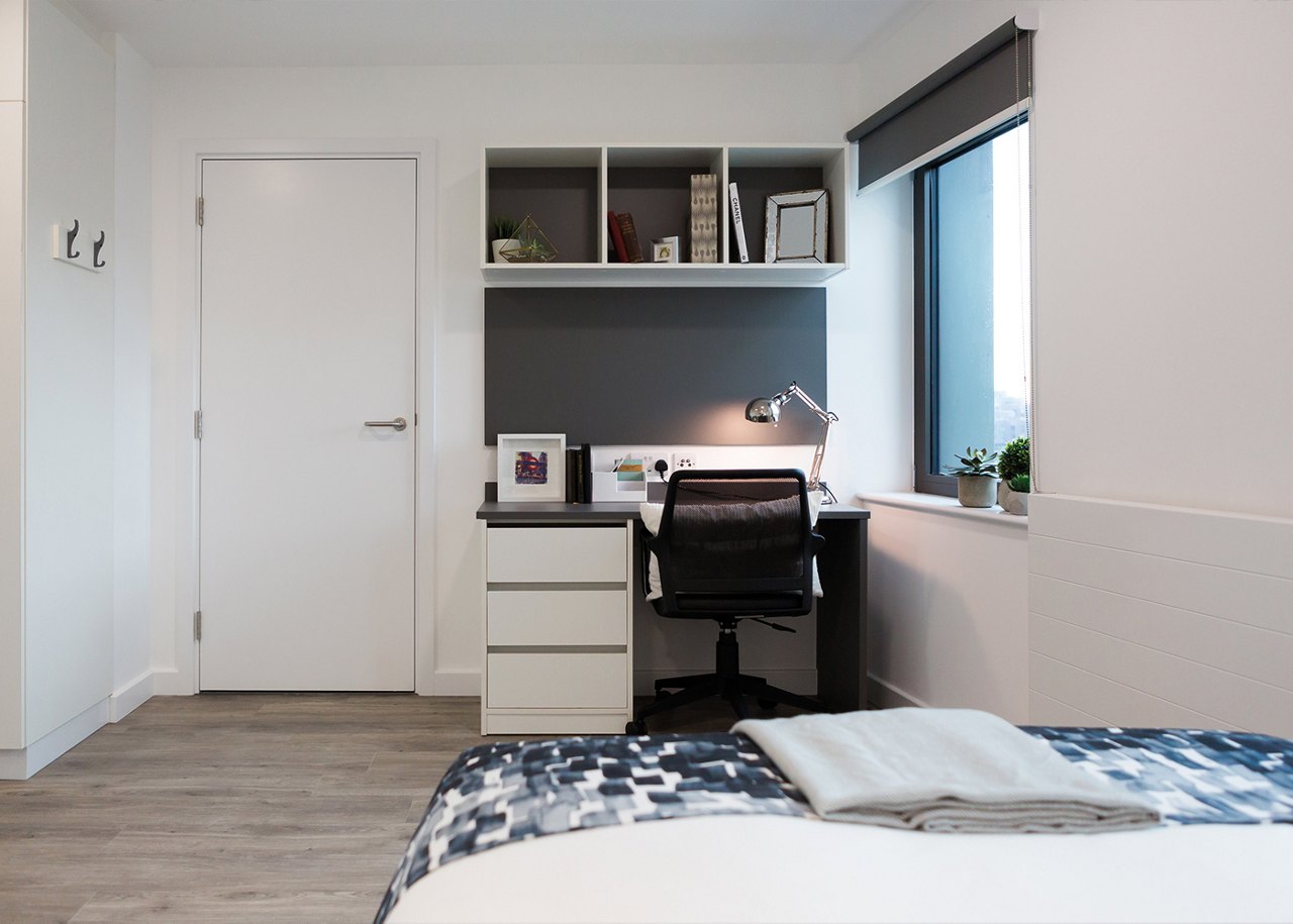 Well-organized student room with a desk, chair, bookshelf, and a bed with a grey and white patterned cover.