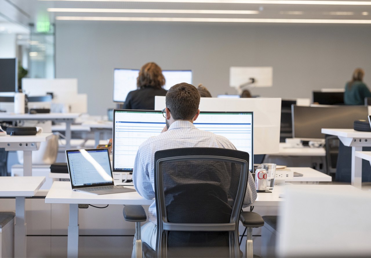 A person working at a desk with dual monitors in a busy open-plan office, focusing on a spreadsheet.