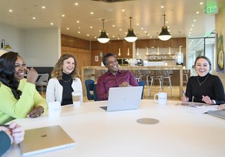 Four diverse professionals smiling and engaging in a lively discussion around a table in a bright, modern office kitchen area.