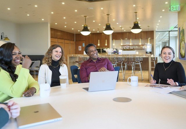 Four diverse professionals smiling and engaging in a lively discussion around a table in a bright, modern office kitchen area.