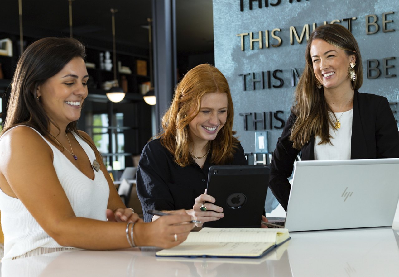 Three businesswomen sharing a cheerful moment around a table with a laptop, tablet, and notebook in a modern office setting.