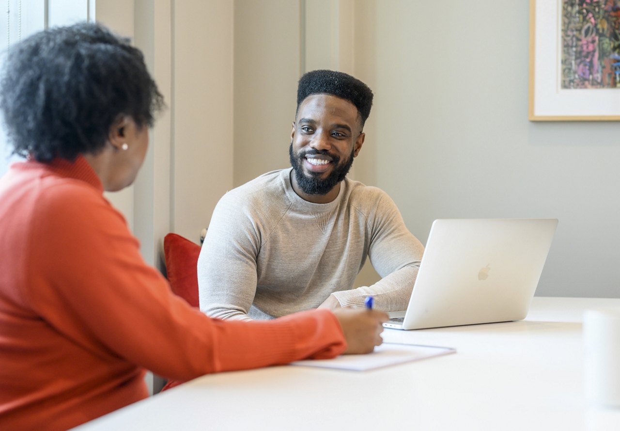 A man with a beard smiling at a colleague during a meeting, with a laptop open and notes being taken in a well-lit office space.