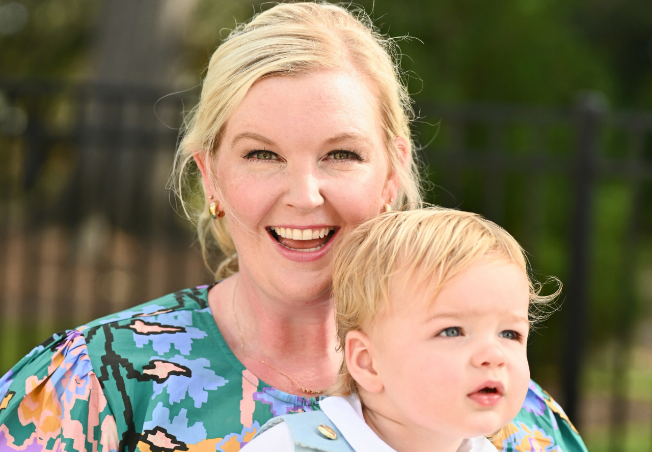 A joyful woman with blonde hair and a colorful blouse holding a toddler outdoors.