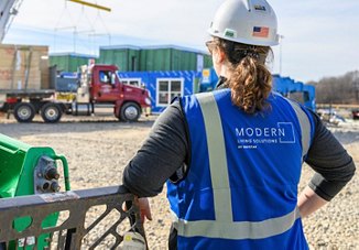 A construction worker in a blue Modern Living Solutions vest by Greystar overlooks a busy construction site with trucks and modular buildings.