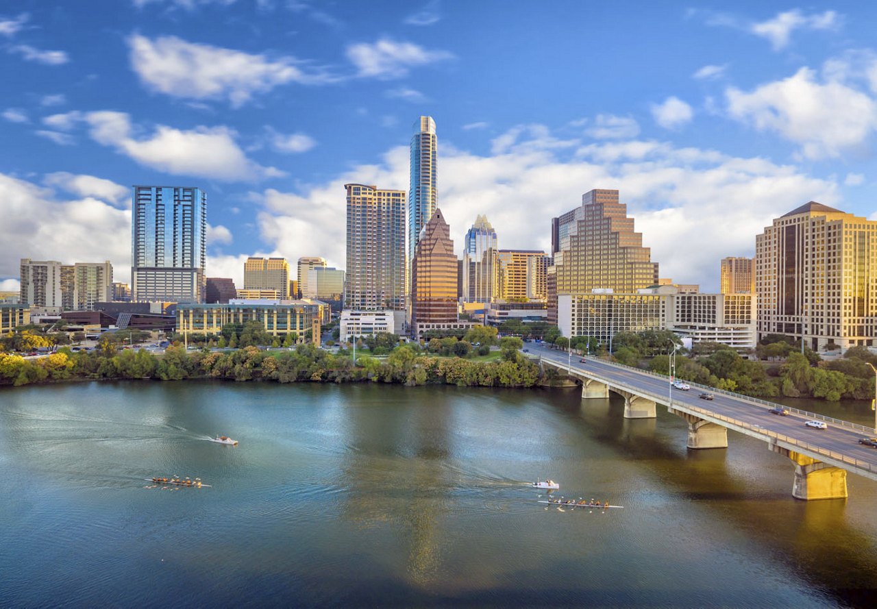 Scenic view of Austin, Texas, with the water and boats passing by, a bridge leading into the city, and a clear blue sky above, highlighting the vibrant cityscape.