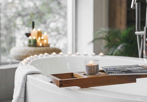 Luxurious bathroom featuring a bathtub surrounded by candles, a tray holding a magazine, and abundant natural light, creating a serene and spa-like atmosphere in a modern apartment.