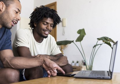 Two African American male roommates sitting at a table in their living room, chatting and looking at a computer, with an indoor plant in the background.