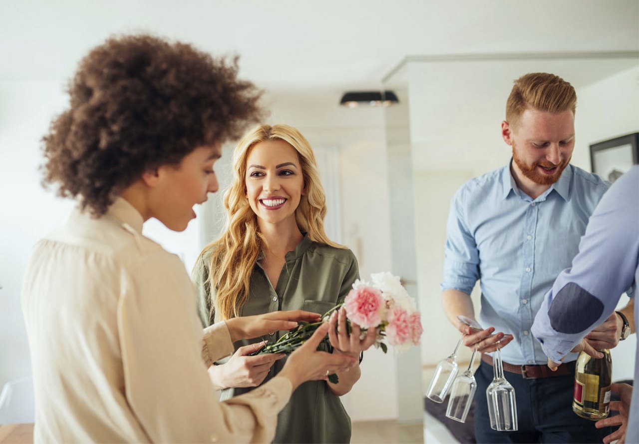 A group of young adults in an apartment warmly greeting guests, holding flowers and wine, and chatting excitedly as they anticipate their visit.