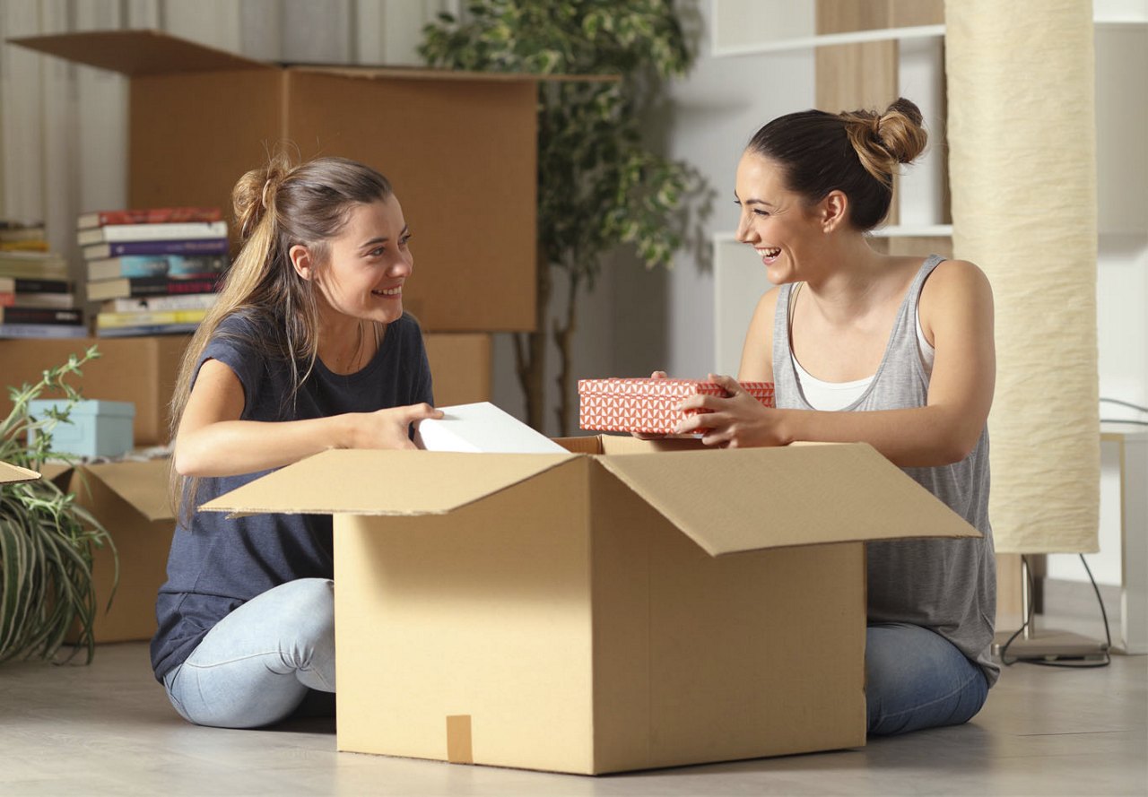 Two female roommates happily unpacking a box together in their apartment, with one moving in and the other excitedly helping, both smiling as they look forward to living together.