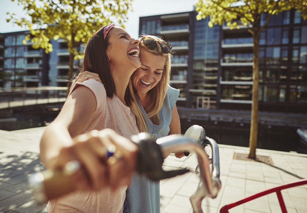  Two females standing together, laughing and holding up a bike, with a building and trees in the background.