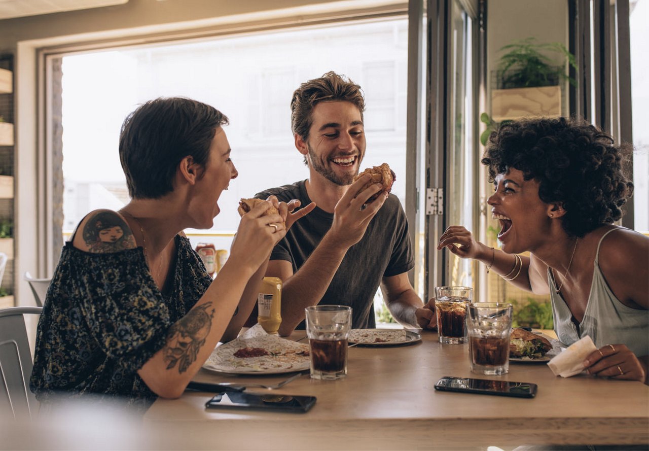 A group of three friends sitting at a table, enjoying a meal together while smiling and laughing, with a large window in the background allowing natural light to fill the space.