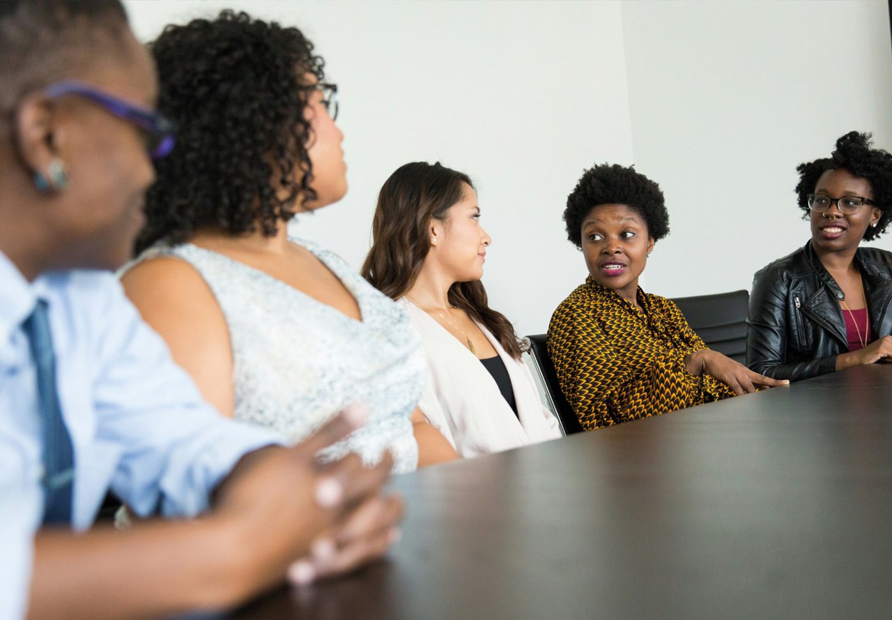 Group of female team members seated together at a long table in a conference room, engaging in a discussion, with a white wall as the backdrop.