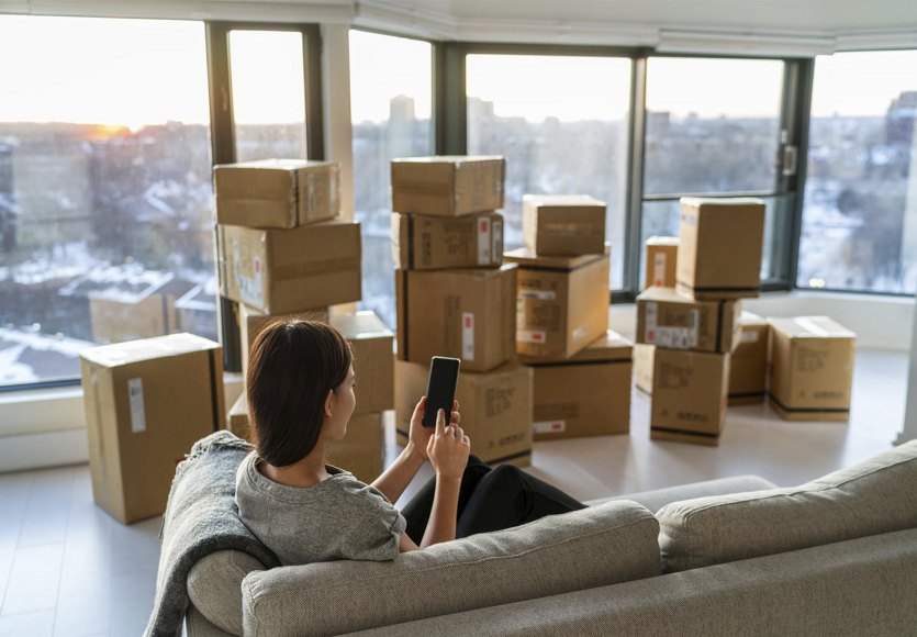 A woman sitting on a couch in her apartment, looking at her phone, surrounded by cardboard boxes ready for moving, with expansive windows in the background offering a view of the city.