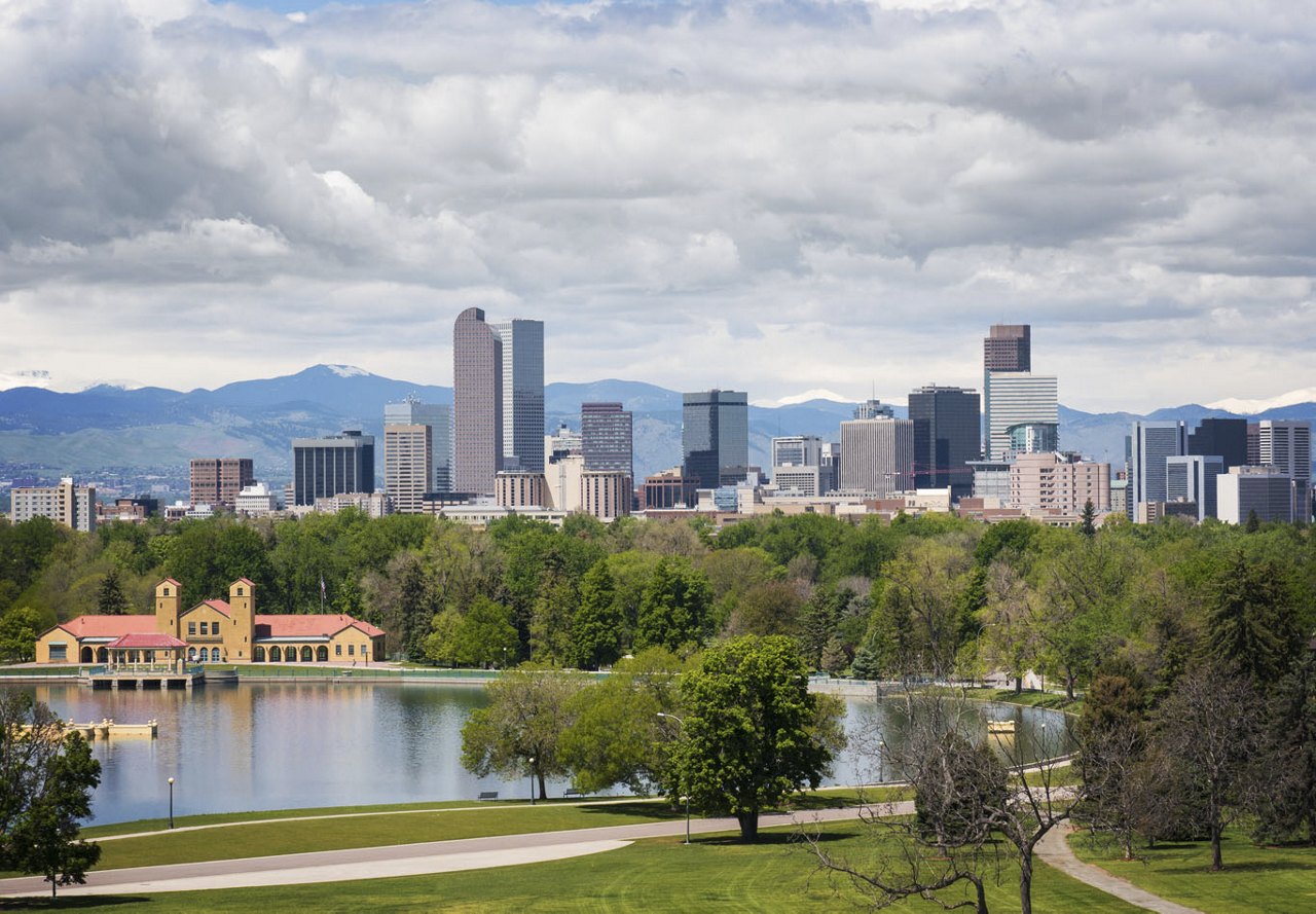 A view of Denver with mountains in the background, a large body of water in the foreground, lush trees surrounding the area, and skyscrapers dotting the city skyline in the distance.