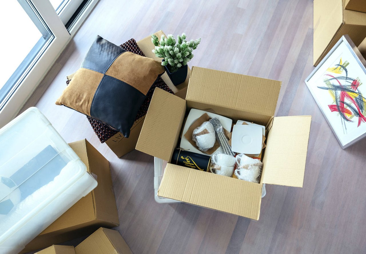 An open cardboard box in the middle of an apartment being packed for a move, filled with mugs and other items, with pillows, plants, and other home decor pieces placed beside the box.