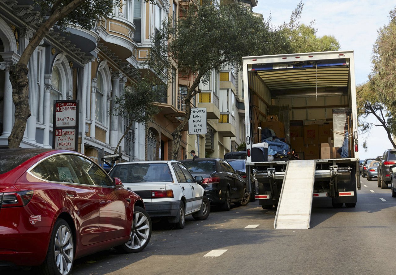 A moving truck parked on a city street with parallel-parked cars alongside it, the ramp extended as it is being loaded with belongings.