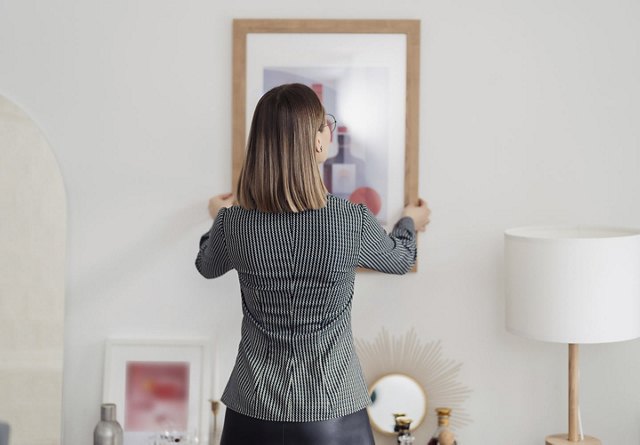 Woman holding a painting in her apartment, ready to hang it on the wall above a decor-styled table.