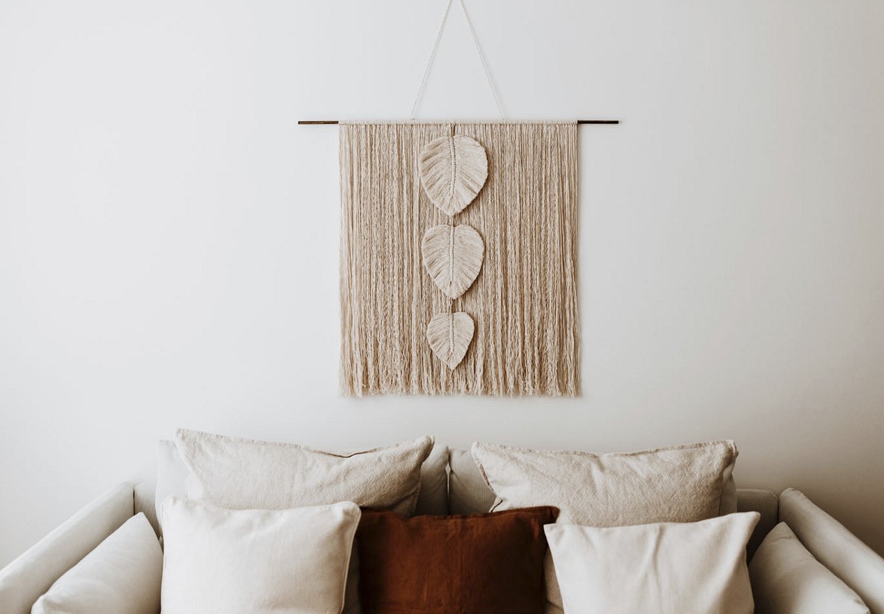 Modern apartment living space featuring a neutral-colored couch with a dark accent pillow, complemented by a woven macrame hanging above the couch for a cozy decorative touch.