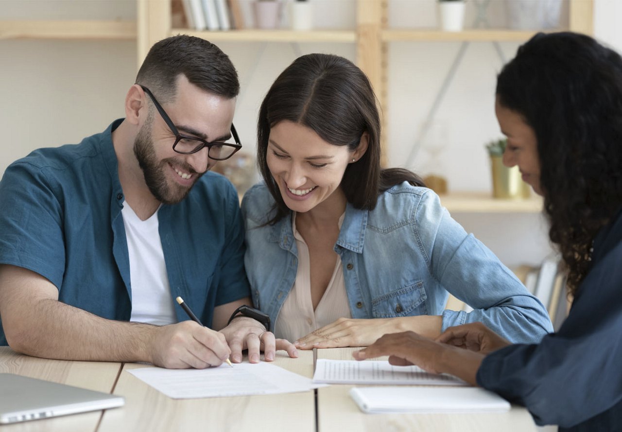 Two people sitting at a table, smiling as they look at a lease document, discussing it with an apartment employee, with a bookcase visible in the background.