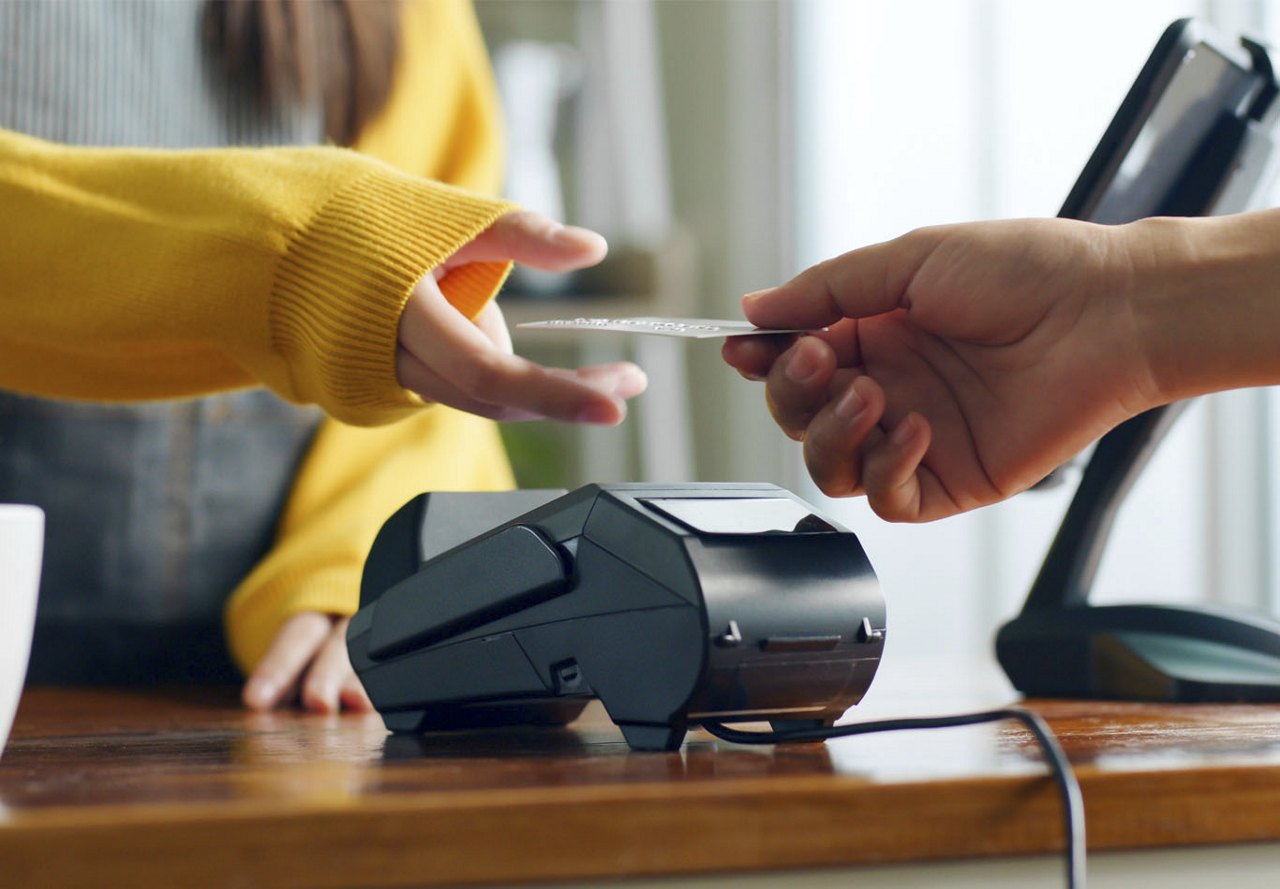 A person handing another person a credit card at a store, with a credit card machine on the desk in front of them as a transaction takes place.