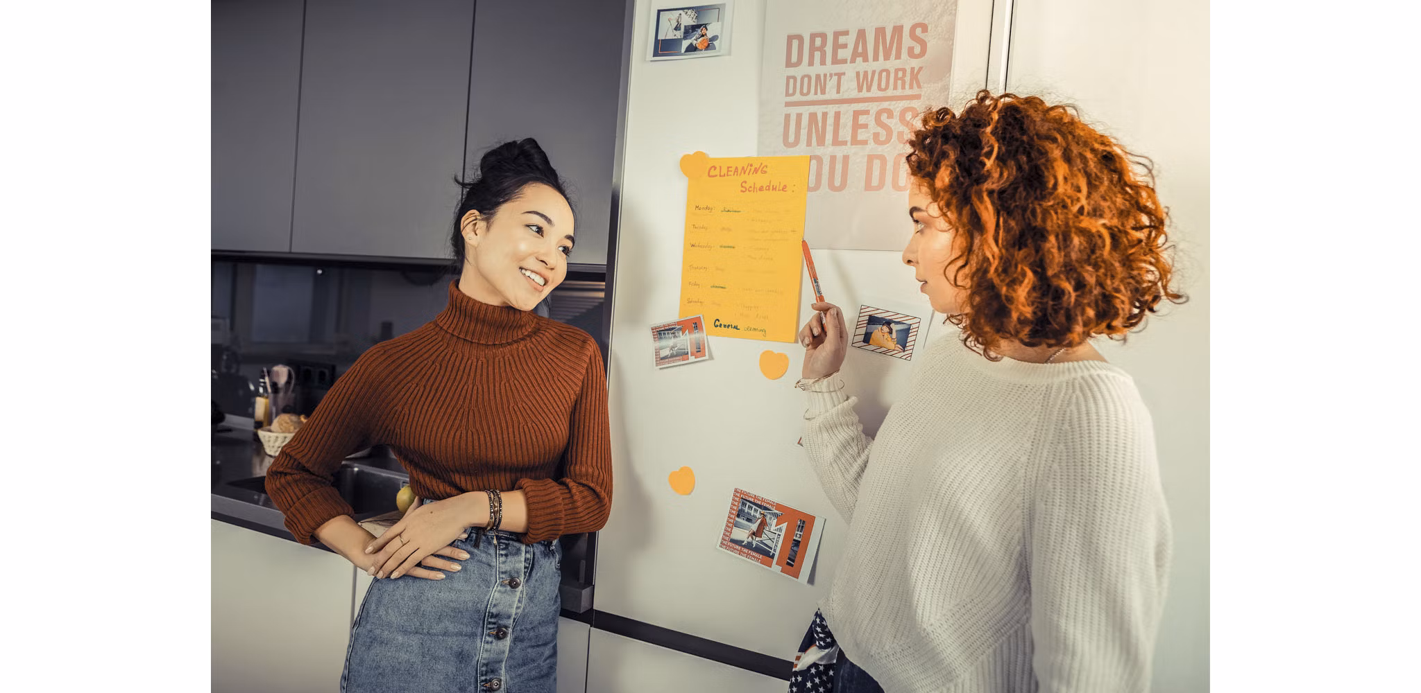  Two female roommates in their apartment kitchen, discussing and pointing at a cleaning schedule on the fridge while planning their household chores together.