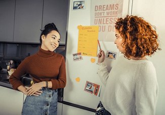  Two female roommates in their apartment kitchen, discussing and pointing at a cleaning schedule on the fridge while planning their household chores together.