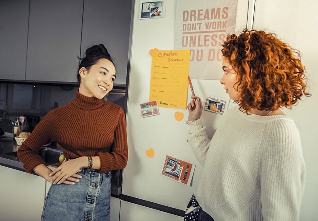  Two female roommates in their apartment kitchen, discussing and pointing at a cleaning schedule on the fridge while planning their household chores together.