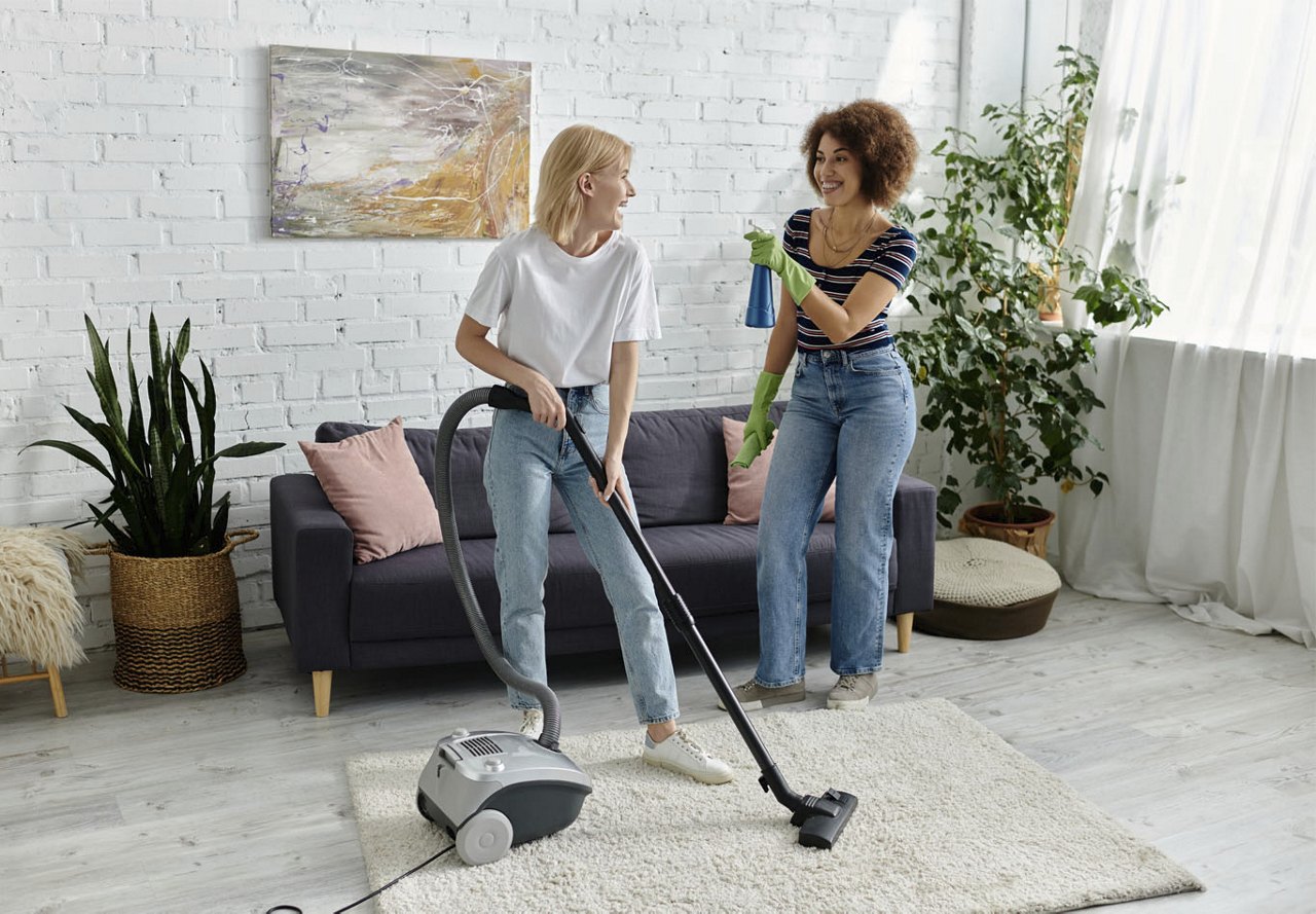 Two female roommates in their living room, laughing as one playfully pretends to spray cleaner at the other, who is vacuuming the carpet, with their stylish home décor visible in the background.