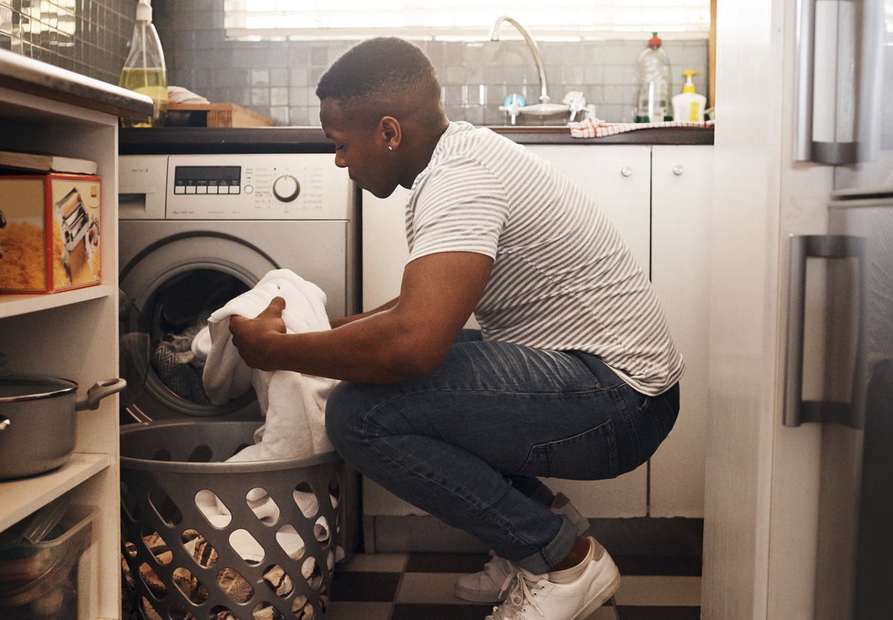 A man holding clothes in front of a washer and dryer in his apartment, transferring them to a basket after finishing his laundry.