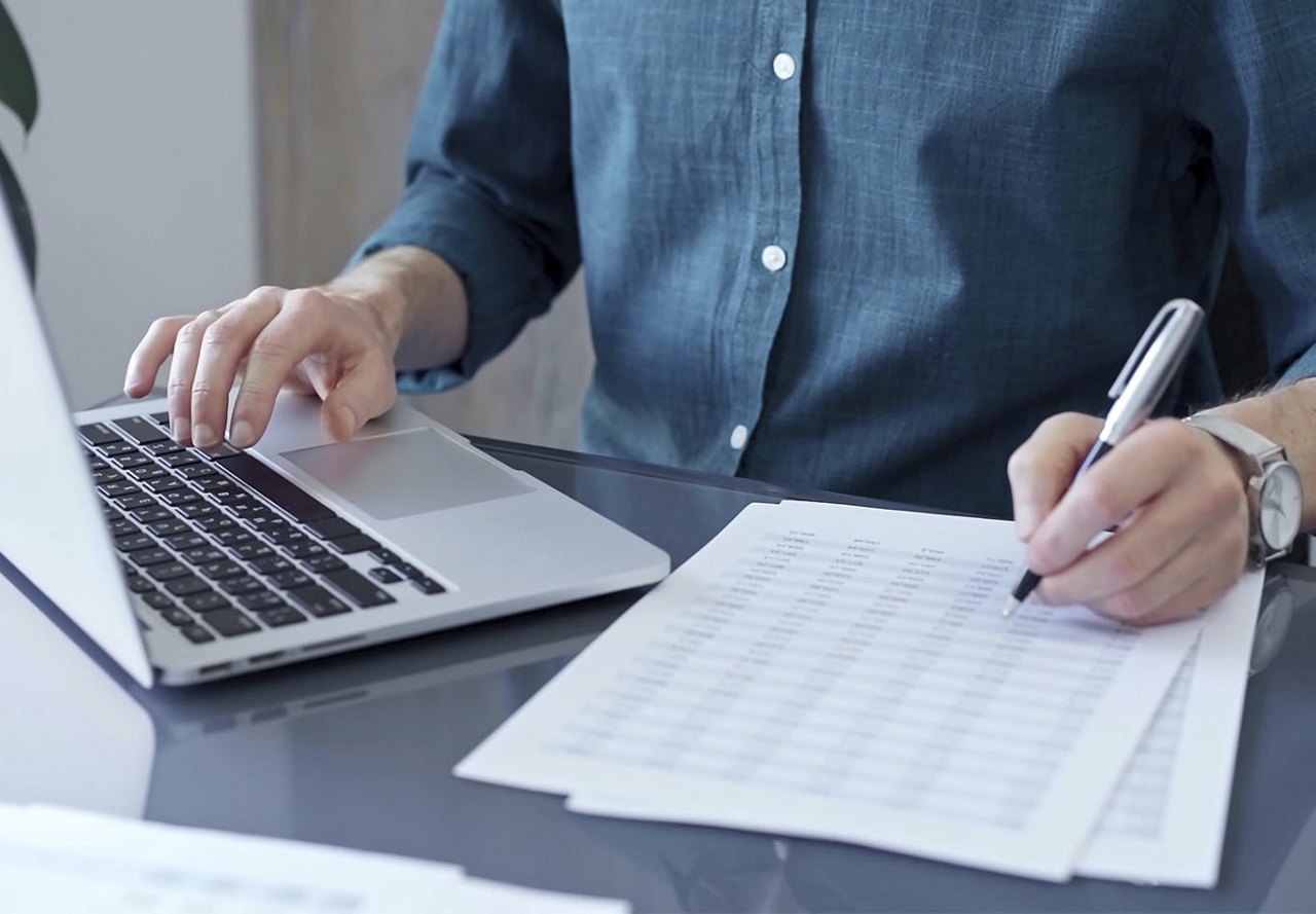 A person sitting at a desk, wearing a denim shirt, looking at a printed spreadsheet while also glancing at their computer, holding a pen to cross off items on the list.