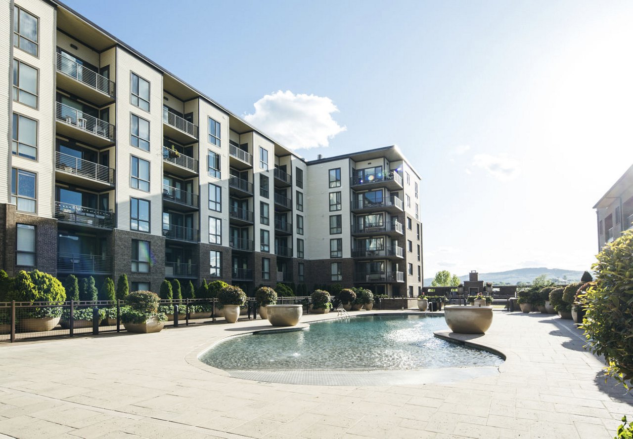 A view of an apartment pool with a modern apartment building behind it, set against a backdrop of a mountain in the distance, surrounded by lush greenery and a clear blue sky.