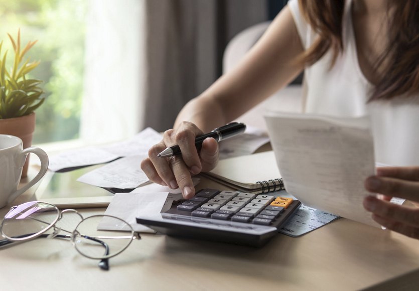 A woman sitting at a table, reviewing a bill and using a calculator to add up expenses, with glasses on the table and a window in the background.