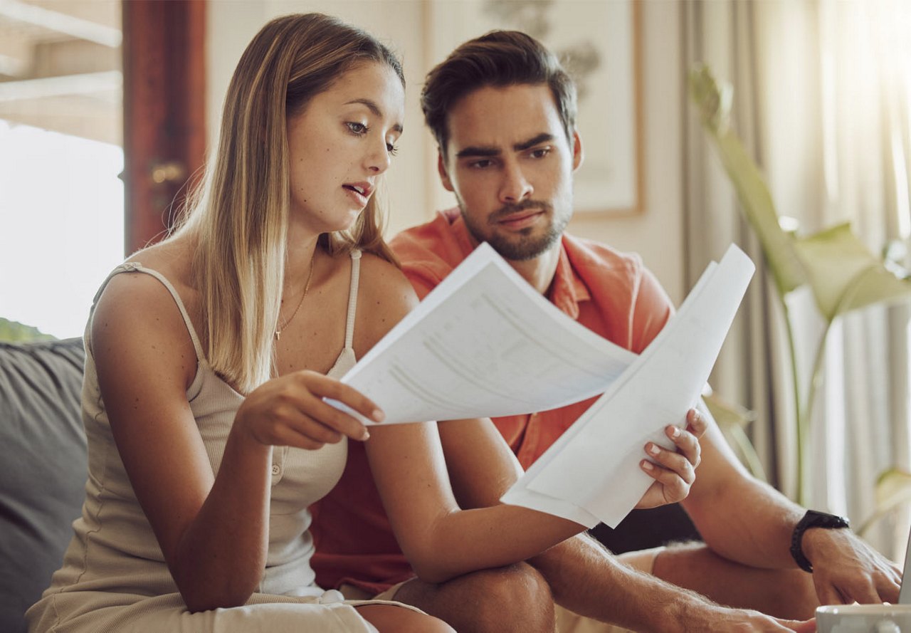 A couple sitting on their apartment couch reviewing papers together as they try to figure out their electric bill, with stylish home decor in the background.